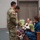 USARIEM COC/Relinquishment of Responsibility 2026 A military ceremony appears to be taking place indoors, likely a promotion or award ceremony. A soldier in Army Combat Uniform (ACU) with a US flag patch is presenting flower bouquets to family members, including two young girls and a woman in a teal dress. Additional military personnel and civilians are seated in the background observing the event.