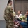 USARIEM COC/Relinquishment of Responsibility 2026 A U.S. Army soldier in OCP (Operational Camouflage Pattern) uniform is receiving flowers from a young girl during what appears to be a military ceremony. A woman in a blue dress, likely the soldier's spouse or family member, is seated nearby holding additional flowers along with a younger child. Other military personnel and civilians are visible seated in the background.