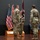 USARIEM COC/Relinquishment of Responsibility 2026 A military ceremony is taking place indoors, likely a change of command or activation ceremony. Three soldiers in the foreground are participating in a guidon (unit flag) transfer, with a maroon/burgundy colored flag being passed between personnel. A fourth soldier stands at attention in the background near the right side of the stage.
