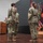 USARIEM COC/Relinquishment of Responsibility 2026 A military ceremony is taking place on a raised wooden stage, likely a change of command or award ceremony. Three uniformed U.S. Army soldiers in OCP (Operational Camouflage Pattern) uniforms are positioned center stage, with one appearing to interact closely with another. A fourth soldier stands at a podium in the background, and multiple military unit flags and the U.S. flag are displayed on a flag stand to the right.