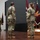 USARIEM COC/Relinquishment of Responsibility 2026 A formal U.S. Army military ceremony is taking place on a stage, likely a change of command or award ceremony. Three soldiers in Army Combat Uniforms (ACUs) are present, with one speaking at a podium and two standing at attention facing the speaker. Military guidons and unit flags are displayed on a wooden stand to the right.