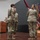 USARIEM COC/Relinquishment of Responsibility 2026 A military ceremony is taking place on a stage, likely a change of command or activation ceremony. Several soldiers in OCP (Operational Camouflage Pattern) uniforms are gathered near a set of unit colors and flags, with one soldier at a podium in the background. The presence of a maroon/burgundy guidon and multiple unit flags suggests a formal military organizational event.