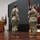 USARIEM COC/Relinquishment of Responsibility 2026 A military ceremony is taking place on a stage, likely a change of command or activation ceremony. Three soldiers in Army Combat Uniforms (ACU/OCP pattern) are present, with one speaking at a podium and two others standing at attention near unit guidons. The setting appears to be an indoor auditorium or meeting hall.