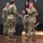 USARIEM COC/Relinquishment of Responsibility 2026 A military ceremony is taking place on a stage, likely a change of command or award ceremony. Two soldiers are shaking hands in the foreground while a third soldier holds a unit guidon/flag in the background. All personnel are wearing US Army Operational Camouflage Pattern (OCP) uniforms.