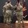 USARIEM COC/Relinquishment of Responsibility 2026 A U.S. Army change of command ceremony is taking place on a stage, with a guidon (unit flag) being passed between soldiers. Three soldiers in Army Combat Uniforms (ACU/OCP pattern) are the central figures, with one soldier transferring the maroon guidon to another. Military flags and a wooden flag stand are visible in the background.
