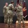 USARIEM COC/Relinquishment of Responsibility 2026 A military change of command ceremony is taking place on a stage, with a soldier passing or receiving a unit guidon (flag) to/from another soldier. Three soldiers in U.S. Army OCP (Operational Camouflage Pattern) uniforms are the central figures, with unit colors and campaign streamers visible in the background. This formal ceremony symbolizes the transfer of command authority within a U.S. Army unit.