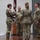 USARIEM COC/Relinquishment of Responsibility 2026 A military ceremony, likely a change of command or activation ceremony, is taking place on a stage. Four soldiers in Army Combat Uniforms (ACU/OCP pattern) are participating, with one soldier passing or receiving a unit guidon (maroon/burgundy flag). Additional flags and military colors are visible in the background.