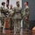 USARIEM COC/Relinquishment of Responsibility 2026 A military ceremony is taking place on a stage or platform, likely a change of command or unit activation/deactivation ceremony. Four U.S. Army soldiers in OCP (Operational Camouflage Pattern) uniforms are participating, with one soldier passing or receiving a unit guidon (flag). Military flags including the U.S. flag and unit colors are displayed on stands in the background.