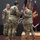 USARIEM COC/Relinquishment of Responsibility 2026 Three U.S. Army soldiers in OCP (Operational Camouflage Pattern) uniforms are participating in a formal military change of command or guidon ceremony on a stage. Two soldiers are in the process of transferring a maroon/burgundy unit guidon (flag), while a third soldier stands at attention observing. Military colors and unit flags are displayed on a stand in the background.