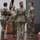 USARIEM COC/Relinquishment of Responsibility 2026 A military ceremony is taking place on a stage or platform, likely a change of command or unit activation/deactivation ceremony. Four soldiers in Army Combat Uniforms (ACU/OCP pattern) are participating in a guidon transfer, with one soldier passing a maroon/burgundy unit guidon to another. Military flags and unit colors are displayed on a stand to the right.