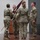 USARIEM COC/Relinquishment of Responsibility 2026 A military change of command or activation ceremony is taking place on an indoor stage. Four soldiers in OCP (Operational Camouflage Pattern) uniforms are participating in a guidon transfer, with one soldier passing a furled maroon/burgundy guidon to another. Military colors and unit flags are visible in the background to the right.