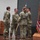 USARIEM COC/Relinquishment of Responsibility 2026 Four U.S. Army soldiers in OCP (Operational Camouflage Pattern) uniforms are participating in a formal military ceremony on a stage. The scene appears to be a change of command or guidon transfer ceremony, with one soldier holding a guidon staff. Unit colors and the U.S. flag are displayed on a flag stand to the right.