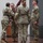 USARIEM COC/Relinquishment of Responsibility 2026 A military ceremony is taking place indoors, likely a change of command or activation/deactivation ceremony. Four soldiers in U.S. Army OCP (Operational Camouflage Pattern) uniforms are participating in a guidon transfer, with one soldier passing a maroon/burgundy guidon to another. A unit colors flag and U.S. flag are displayed on a wooden stand in the background.