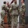 USARIEM COC/Relinquishment of Responsibility 2026 A U.S. Army military ceremony is taking place on a raised wooden stage, likely a change of command or activation/deactivation ceremony. Four soldiers in OCP (Operational Camouflage Pattern) uniforms are engaged in the passing of a guidon (maroon/burgundy unit flag). Military colors including the U.S. flag and a unit flag are displayed on a stand to the right.