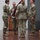 USARIEM COC/Relinquishment of Responsibility 2026 A military ceremony is taking place on a stage or platform, likely a change of command or unit activation/deactivation ceremony. Four U.S. Army soldiers in OCP (Operational Camouflage Pattern) uniforms are gathered around a furled guidon or unit colors draped in maroon. Flags including the U.S. flag and a maroon unit guidon are displayed on stands in the background.