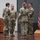 USARIEM COC/Relinquishment of Responsibility 2026 Four U.S. Army soldiers in MultiCam/OCP camouflage uniforms are gathered on a wooden stage during what appears to be a military ceremony. They are engaged in conversation or a formal interaction, with unit colors and an American flag displayed on a stand to the right. A wooden podium with a microphone is visible in the background.