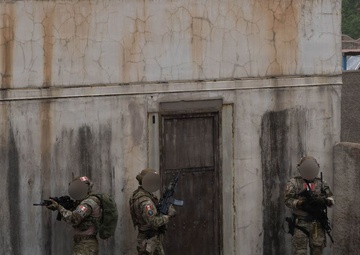 Three armed personnel in military/tactical uniforms are conducting what appears to be a building clearance or close-quarters training exercise along a weathered concrete wall with a door. The individuals are positioned in a tactical stack formation, with weapons raised, approaching a doorway. A bicycle is leaning against the wall to the left, and the setting resembles a military training facility or mock urban environment.