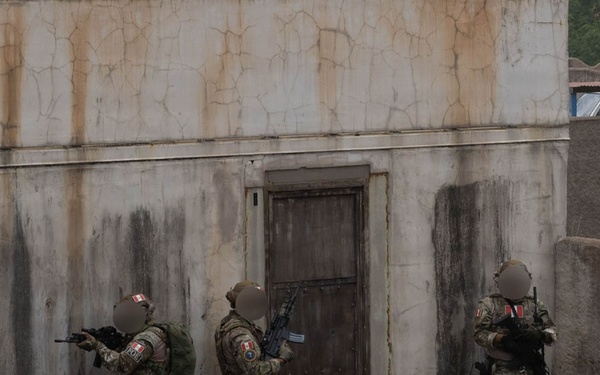 Three armed personnel in military/tactical uniforms are conducting what appears to be a building clearance or close-quarters training exercise along a weathered concrete wall with a door. The individuals are positioned in a tactical stack formation, with weapons raised, approaching a doorway. A bicycle is leaning against the wall to the left, and the setting resembles a military training facility or mock urban environment.