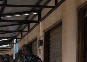 A group of armed military personnel in camouflage uniforms are conducting a tactical building clearance or entry drill along a covered walkway adjacent to a concrete structure with a roll-up metal shutter door. The lead operator is pressing against the wall in a stacked formation, aiming a rifle equipped with an optic and an underbarrel launcher. At least one personnel visible in the background displays what appears to be a Philippine flag patch on their sleeve.