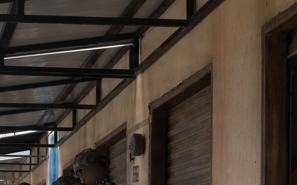 A group of armed military personnel in camouflage uniforms are conducting a tactical building clearance or entry drill along a covered walkway adjacent to a concrete structure with a roll-up metal shutter door. The lead operator is pressing against the wall in a stacked formation, aiming a rifle equipped with an optic and an underbarrel launcher. At least one personnel visible in the background displays what appears to be a Philippine flag patch on their sleeve.
