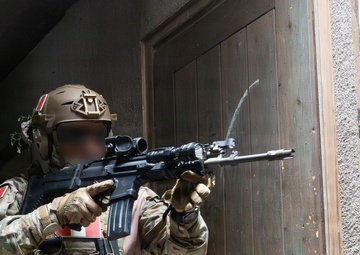 A heavily equipped special operations soldier in multicam camouflage is conducting a tactical entry or building clearance operation near a wooden door. The operator is aiming a rifle with an optic and tactical light toward a doorway. A second operator is partially visible in the background, and the soldier displays visible unit patches including what appears to be a national flag patch.