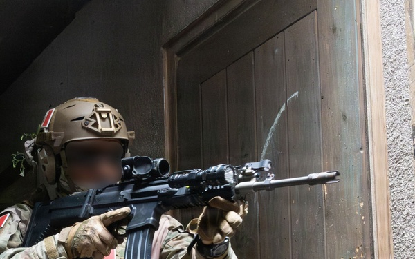 A heavily equipped special operations soldier in multicam camouflage is conducting a tactical entry or building clearance operation near a wooden door. The operator is aiming a rifle with an optic and tactical light toward a doorway. A second operator is partially visible in the background, and the soldier displays visible unit patches including what appears to be a national flag patch.