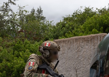 A heavily equipped special operations soldier wearing Peruvian flag patches is in a tactical kneeling position behind a vehicle, holding an assault rifle. The operator's face has been digitally blurred, suggesting this is an official or semi-official military/law enforcement photograph. The setting appears to be a training environment with a concrete wall and vegetation in the background.