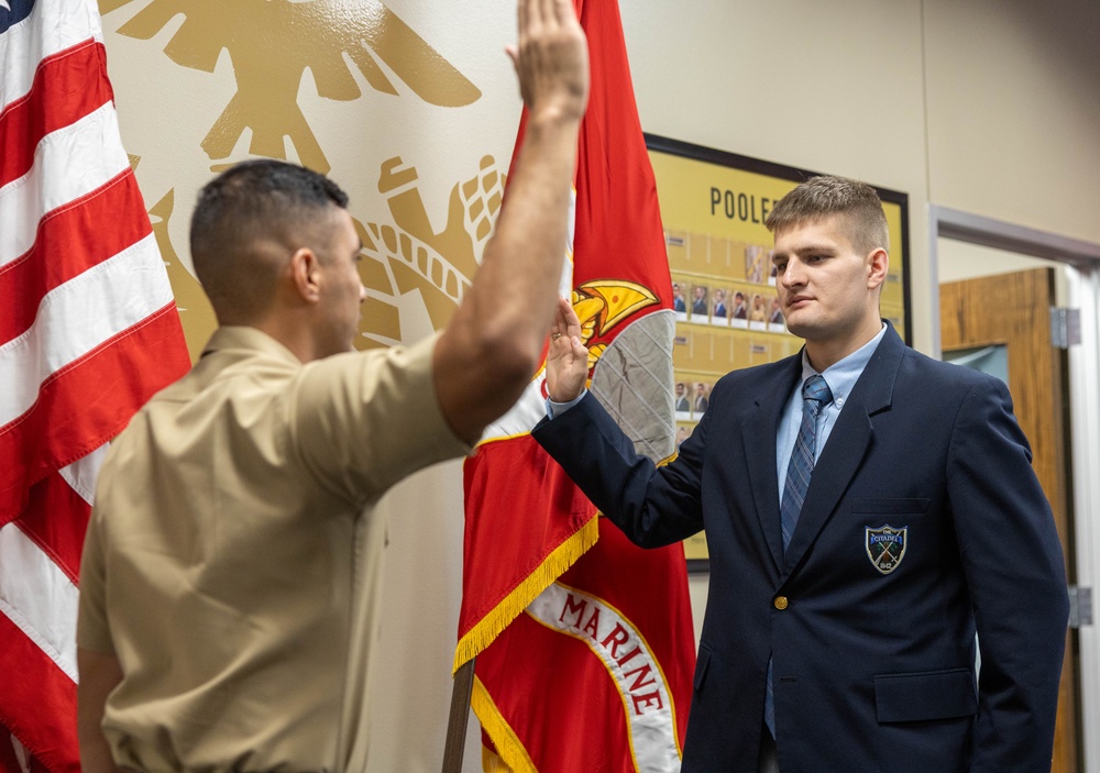 A military enlistment or oath-of-office ceremony is taking place at what appears to be a Marine Corps recruiting station. A Marine in service uniform administers an oath to a young civilian man in a navy blazer, both with right hands raised. The setting includes American and Marine Corps flags, and a 'Poolee' board visible on the wall in the background.