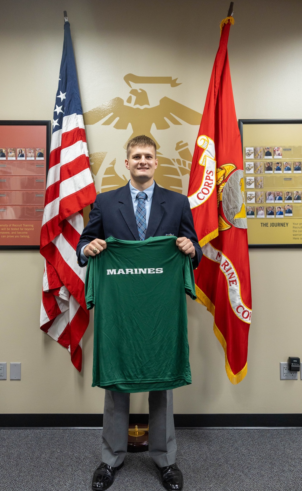 A young man in a dark blazer and gray slacks stands in what appears to be a U.S. Marine Corps recruiting office, holding up a green 'MARINES' jersey. He is positioned between an American flag and a Marine Corps flag, with a large Marine Corps Eagle, Globe, and Anchor emblem visible on the wall behind him. The setting and jersey suggest this is an enlistment or signing ceremony.