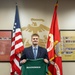 A young man in a dark blazer and gray slacks stands in what appears to be a U.S. Marine Corps recruiting office, holding up a green 'MARINES' jersey. He is positioned between an American flag and a Marine Corps flag, with a large Marine Corps Eagle, Globe, and Anchor emblem visible on the wall behind him. The setting and jersey suggest this is an enlistment or signing ceremony.
