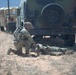 A soldier in full combat gear and camouflage uniform is kneeling beside a downed person on arid desert terrain, appearing to perform a casualty assessment or medical aid. The scene takes place near the rear of a camouflage-painted HMMWV (Humvee) military vehicle, with additional Humvees visible in the background. The setting and equipment suggest a military training exercise in a desert environment.