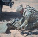 A soldier in MultiCam/OCP camouflage uniform is kneeling on arid desert terrain, appearing to administer first aid or treat a downed fellow soldier. The scene takes place near a military vehicle, with a rifle and equipment visible on the ground. The setting suggests a field training exercise or combat scenario in a desert environment.