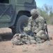 A soldier in OCP (Operational Camouflage Pattern) uniform is kneeling beside a downed fellow soldier on arid, sandy terrain next to a military HMMWV (Humvee). The standing soldier appears to be performing tactical combat casualty care (TCCC) or a medical assessment on the prone individual. The setting and equipment suggest a training exercise or field operation in a desert environment.