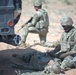 The image shows military personnel in MultiCam/OCP camouflage uniforms conducting what appears to be a combat casualty care (TCCC) training exercise in an arid, desert-like environment. One soldier is kneeling beside a prone soldier on the ground, performing medical aid, while a second soldier maintains a security posture near a military HMMWV (Humvee). The setting and controlled nature of the scenario suggest a field training exercise rather than an actual combat situation.
