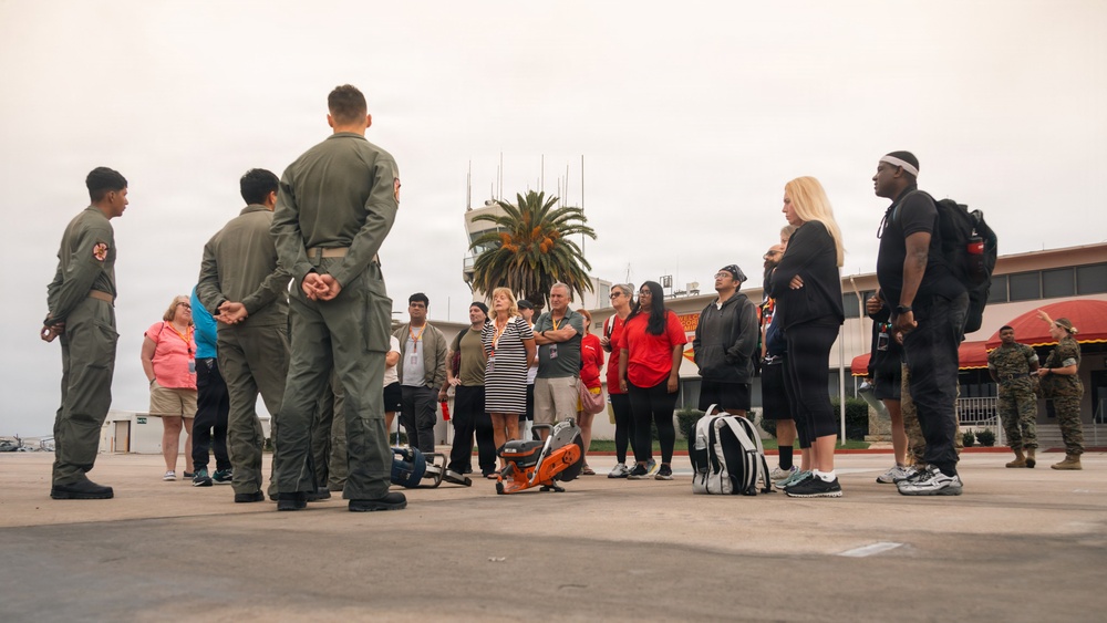 A group of military personnel in olive/green flight suits are briefing or addressing a group of civilians on what appears to be a military airfield tarmac. The setting includes a control tower, palm tree, and a building with a red awning visible in the background. Additional uniformed personnel, likely Marines in camouflage, are visible on the right side near the building.