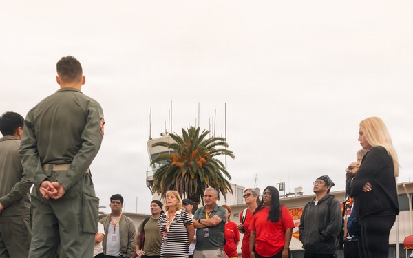 A group of military personnel in olive/green flight suits are briefing or addressing a group of civilians on what appears to be a military airfield tarmac. The setting includes a control tower, palm tree, and a building with a red awning visible in the background. Additional uniformed personnel, likely Marines in camouflage, are visible on the right side near the building.