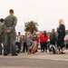 A group of military personnel in olive/green flight suits are briefing or addressing a group of civilians on what appears to be a military airfield tarmac. The setting includes a control tower, palm tree, and a building with a red awning visible in the background. Additional uniformed personnel, likely Marines in camouflage, are visible on the right side near the building.