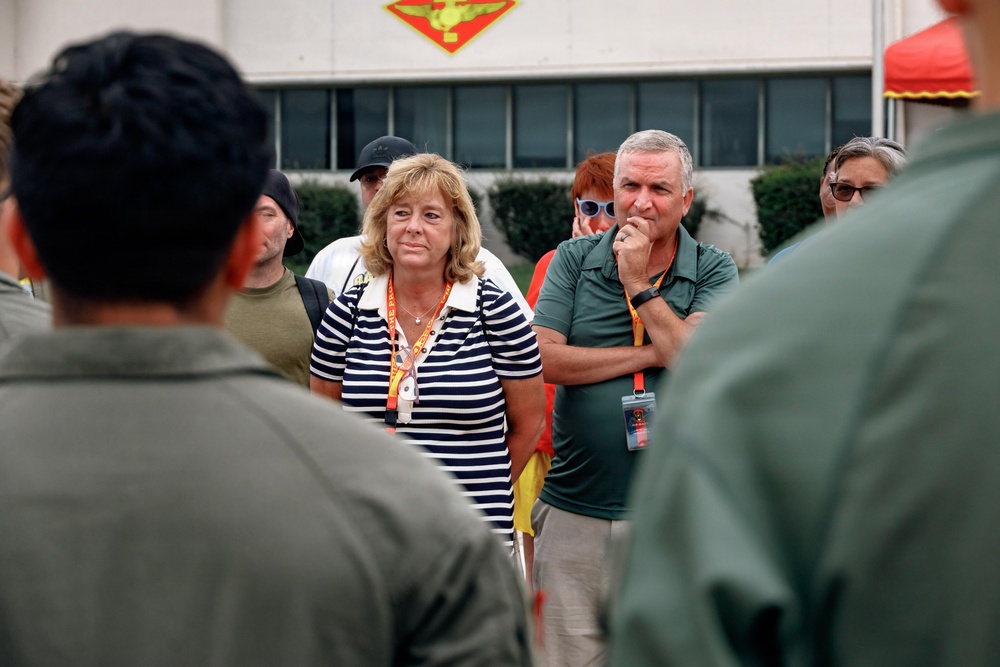 A group of civilians wearing orange/red lanyards with ID badges are gathered outdoors, appearing to listen to a briefing from individuals in military flight suits. The setting appears to be a military installation, evidenced by a unit insignia/emblem visible on the building in the background. The civilians appear attentive and engaged, suggesting this may be a family day, base tour, or official visit event.