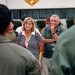 A group of civilians wearing orange/red lanyards with ID badges are gathered outdoors, appearing to listen to a briefing from individuals in military flight suits. The setting appears to be a military installation, evidenced by a unit insignia/emblem visible on the building in the background. The civilians appear attentive and engaged, suggesting this may be a family day, base tour, or official visit event.
