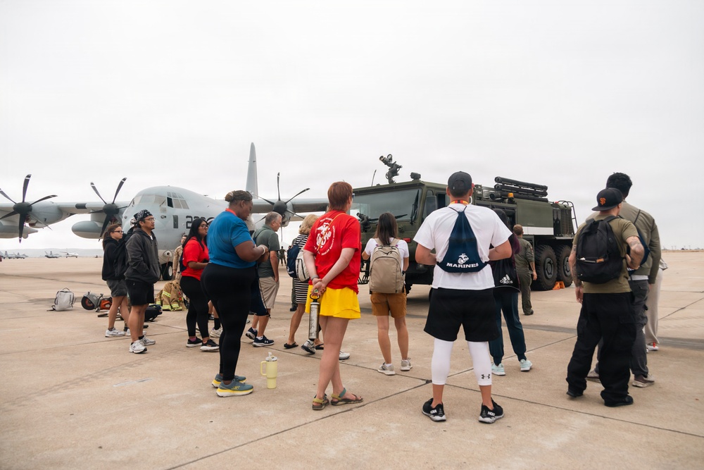 A group of civilians is gathered on a military airfield tarmac for what appears to be a base tour or open house event. They are standing near a gray C-130 Hercules transport aircraft and a large military tactical vehicle. Several visitors are wearing Marines-branded apparel, suggesting this is a Marine Corps installation event.