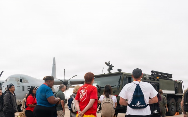 A group of civilians is gathered on a military airfield tarmac for what appears to be a base tour or open house event. They are standing near a gray C-130 Hercules transport aircraft and a large military tactical vehicle. Several visitors are wearing Marines-branded apparel, suggesting this is a Marine Corps installation event.