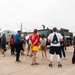 A group of civilians is gathered on a military airfield tarmac for what appears to be a base tour or open house event. They are standing near a gray C-130 Hercules transport aircraft and a large military tactical vehicle. Several visitors are wearing Marines-branded apparel, suggesting this is a Marine Corps installation event.