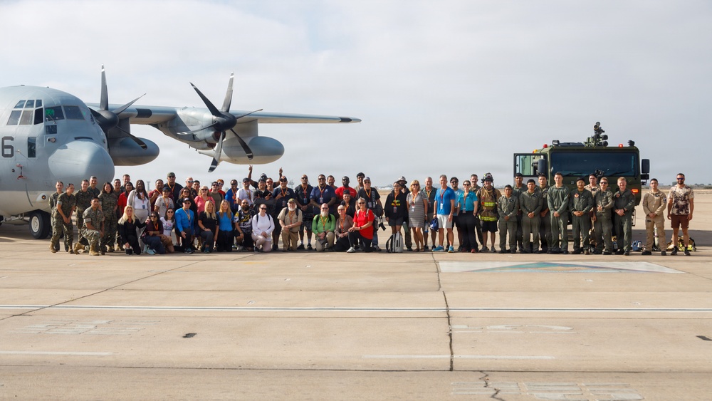 A large group of approximately 50-60 people pose for a group photograph on a military airfield tarmac. The group includes a mix of military personnel in uniform and civilians in casual attire, gathered between a military transport aircraft and a large military vehicle. This appears to be an organized base tour, media day, or community outreach event at a U.S. Marine Corps or Navy air installation.
