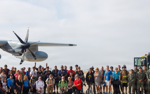 A large group of approximately 50-60 people pose for a group photograph on a military airfield tarmac. The group includes a mix of military personnel in uniform and civilians in casual attire, gathered between a military transport aircraft and a large military vehicle. This appears to be an organized base tour, media day, or community outreach event at a U.S. Marine Corps or Navy air installation.