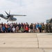 A large group of approximately 50-60 people pose for a group photograph on a military airfield tarmac. The group includes a mix of military personnel in uniform and civilians in casual attire, gathered between a military transport aircraft and a large military vehicle. This appears to be an organized base tour, media day, or community outreach event at a U.S. Marine Corps or Navy air installation.
