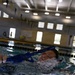 A person is swimming in an indoor pool while using a blue snorkel. The camera angle is at water level, providing a close-up perspective of the swimmer's face and the snorkel equipment. The setting appears to be a large indoor aquatic facility with lane dividers and overhead fluorescent lighting.