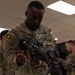 A U.S. Army soldier named Randolph, wearing OCP (Operational Camouflage Pattern) uniform with a '44' patch on the left sleeve, is examining or handling a weapon at what appears to be an indoor equipment demonstration or training event. A bipod-mounted weapon is visible on a white table in the foreground. Other soldiers in uniform are visible in the background.