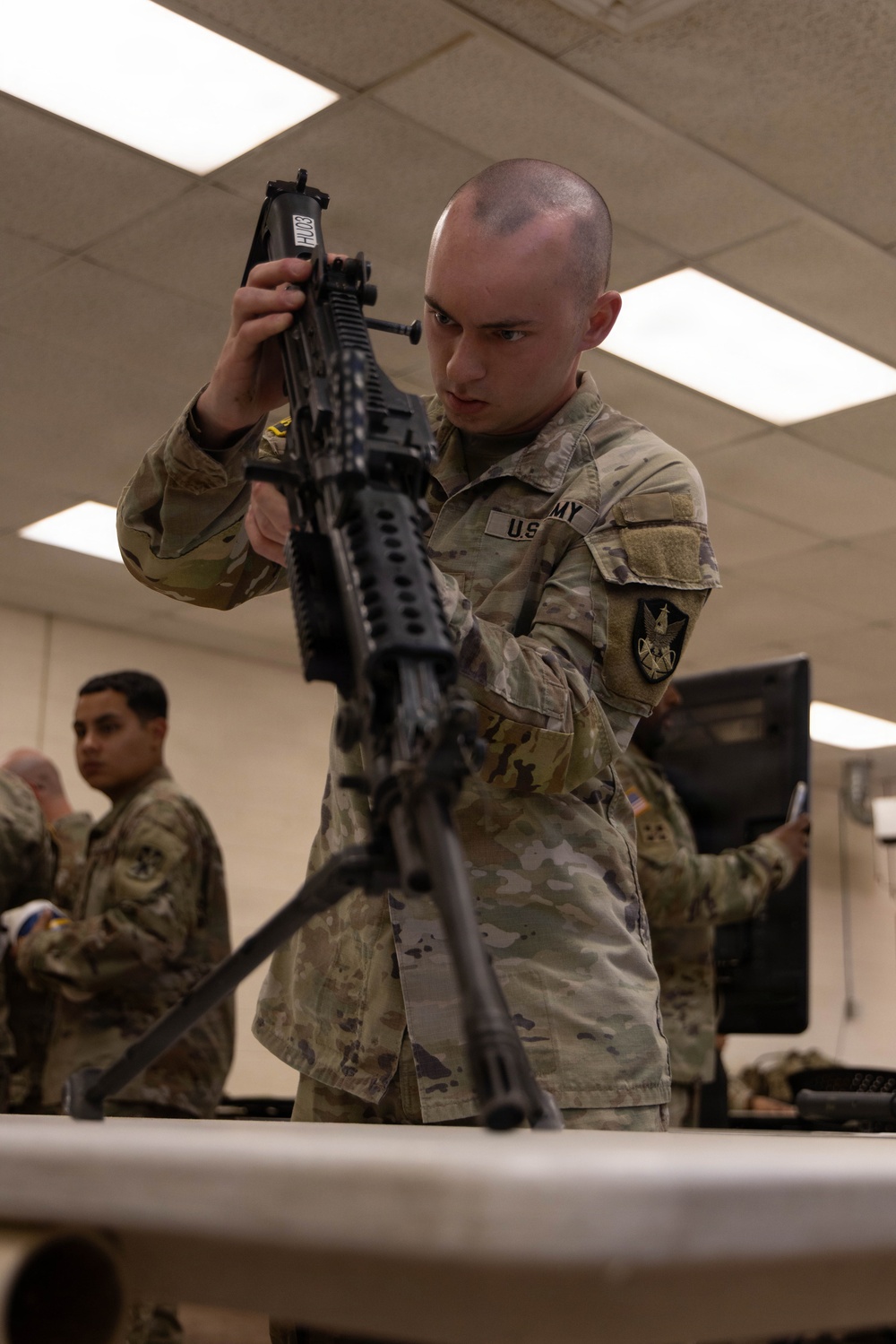 A U.S. Army soldier in OCP (Operational Camouflage Pattern) uniform is inspecting or performing maintenance on what appears to be an M249 SAW (Squad Automatic Weapon) or similar light machine gun mounted on a bipod on a table. The activity appears to take place in an indoor classroom or armory setting. Additional soldiers in the same uniform are visible in the background.