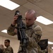 A U.S. Army soldier in OCP (Operational Camouflage Pattern) uniform is inspecting or performing maintenance on what appears to be an M249 SAW (Squad Automatic Weapon) or similar light machine gun mounted on a bipod on a table. The activity appears to take place in an indoor classroom or armory setting. Additional soldiers in the same uniform are visible in the background.