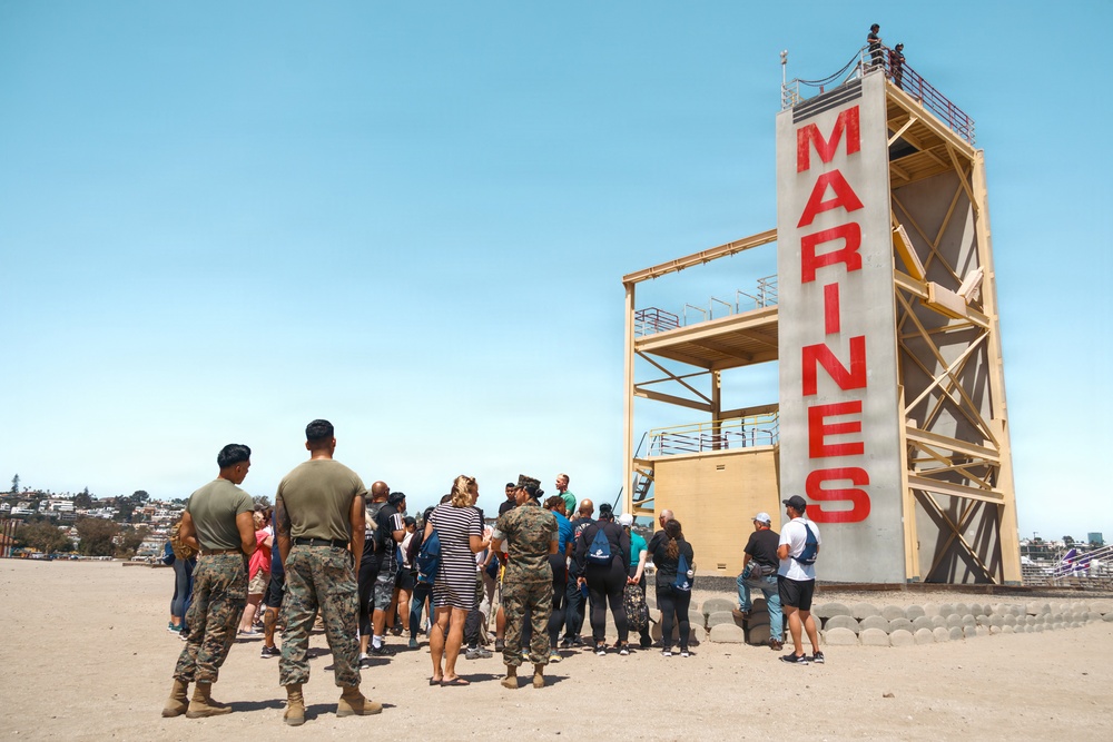 A group of civilians and U.S. Marines gather at the base of a tall training tower marked with the word 'MARINES' in large red letters on a sandy beach. The scene appears to be a public outreach or recruitment event, with military personnel in camouflage uniforms interacting with visitors. The setting is a sunny coastal location with a residential hillside visible in the background.